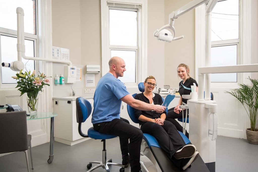 a smiling male and female dentist in a dental clinic showing a patient in dental chair the results of dental work using a mirror