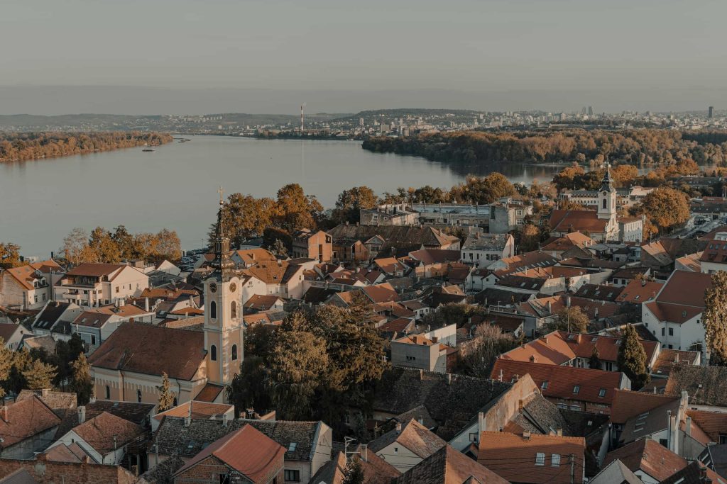 Aerial view over Zemun, Belgrade, showing red-tiled rooftops, a slender church steeple, and the wide Danube River against a distant cityscape