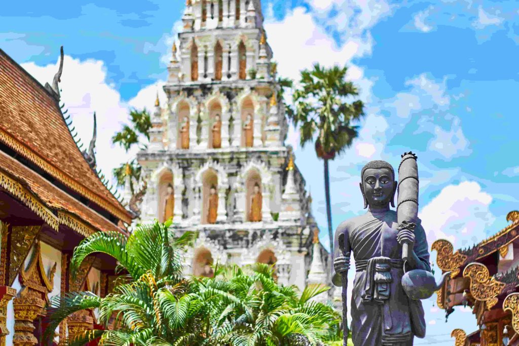 Ornate Buddhist temple complex with palm trees and a standing statue against a bright blue sky.
