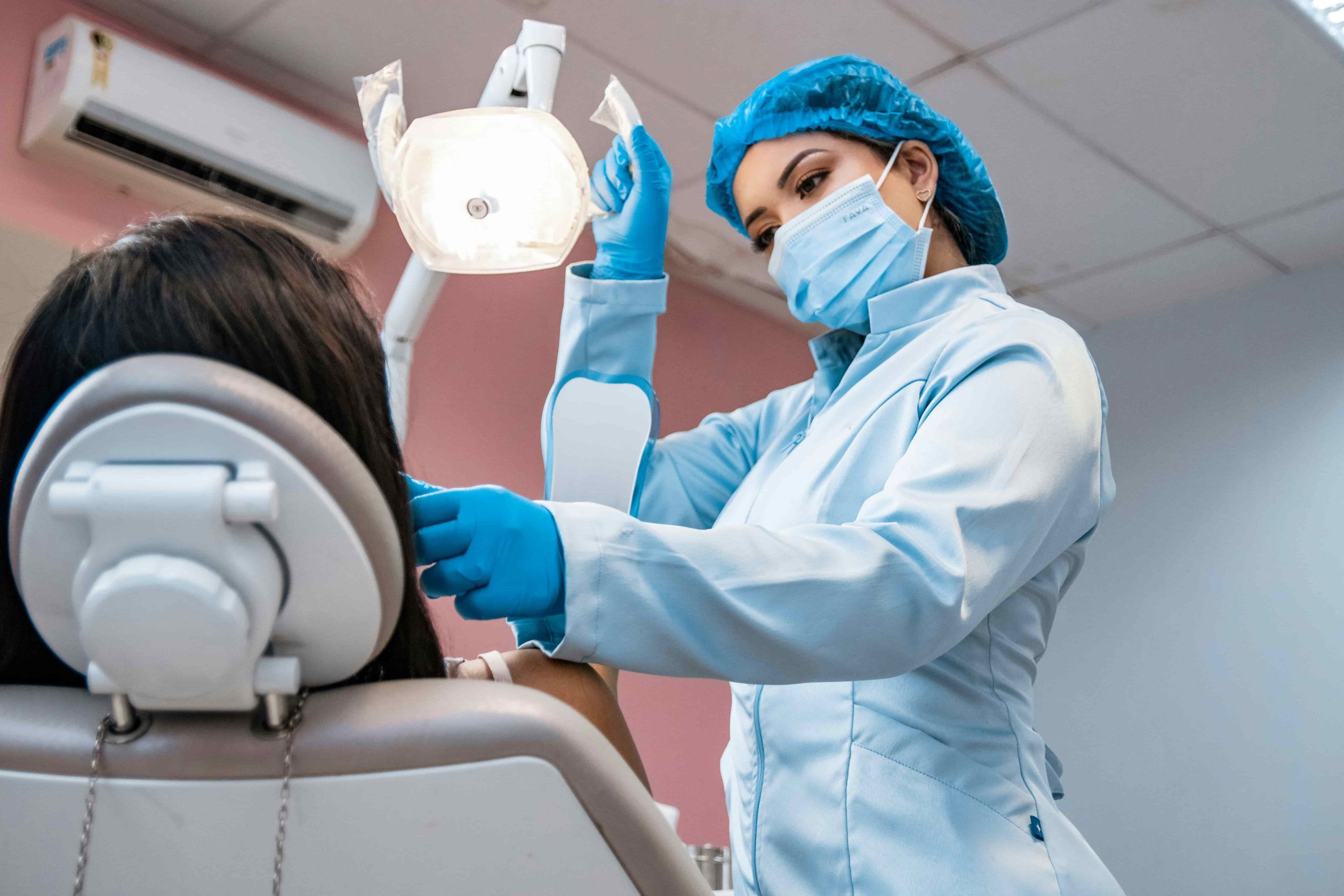 A dentist in a mask, gloves, and surgical cap leans over a patient in a reclined dental chair, adjusting instruments under a bright overhead exam light in a clinic