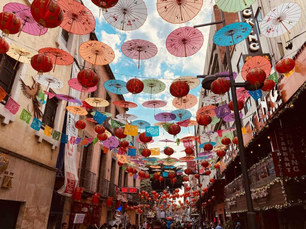 Colorful paper umbrellas and red lanterns strung above a busy city street during a daytime festival.