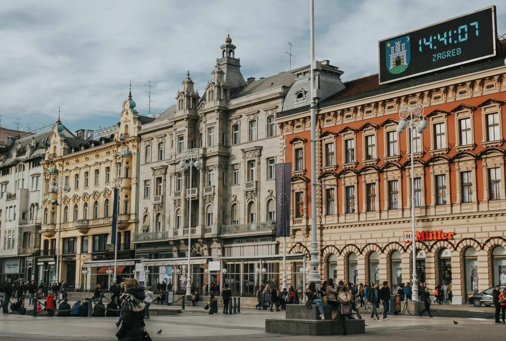 Historic façades lining Zagreb’s Ban Jelačić Square, with pedestrians in the plaza and a large digital clock displaying the city’s coat of arms