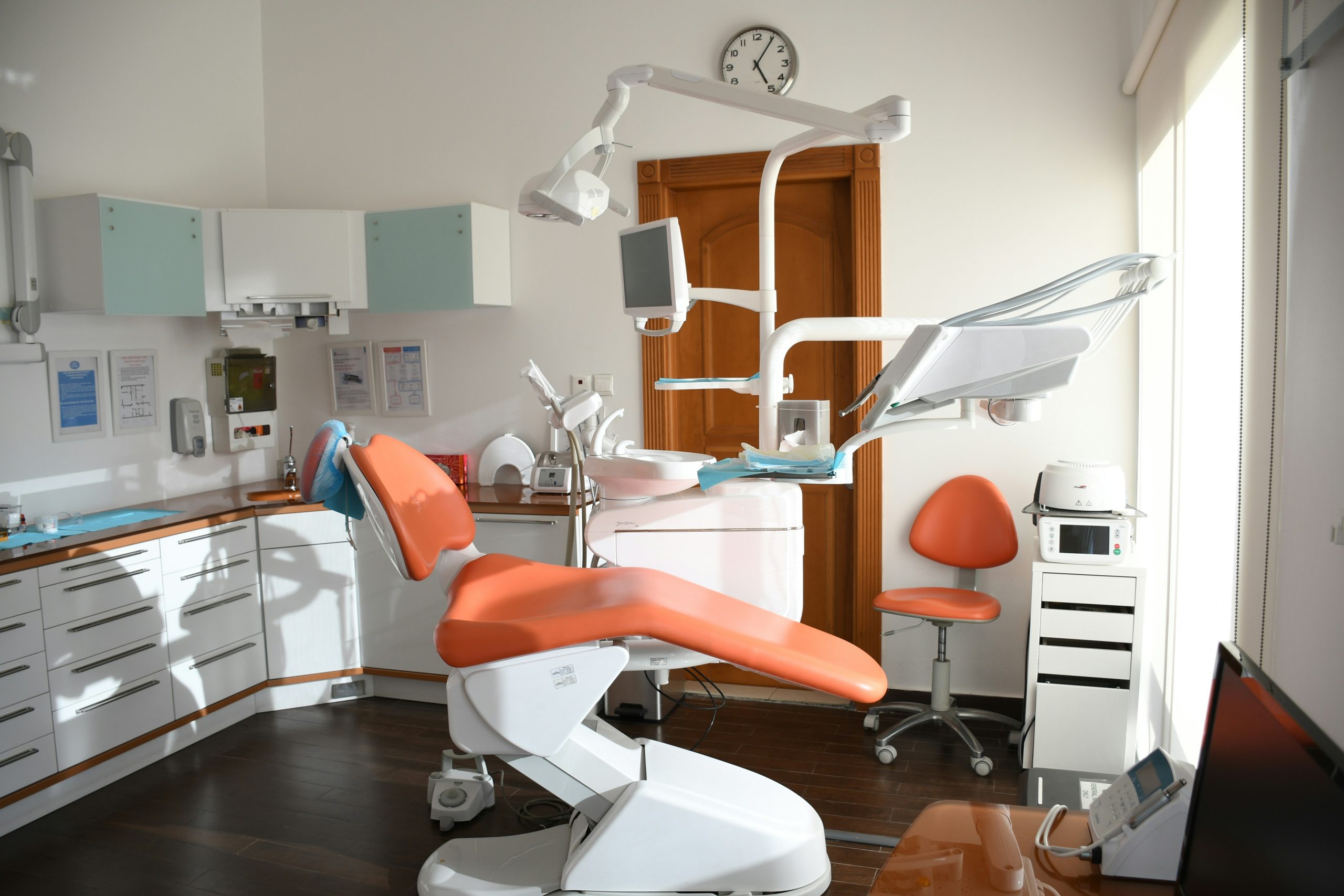 Modern dental clinic interior with an orange dental chair, white cabinetry, a large wall clock, and professional dental equipment, ideal for a dental practice in a city setting.